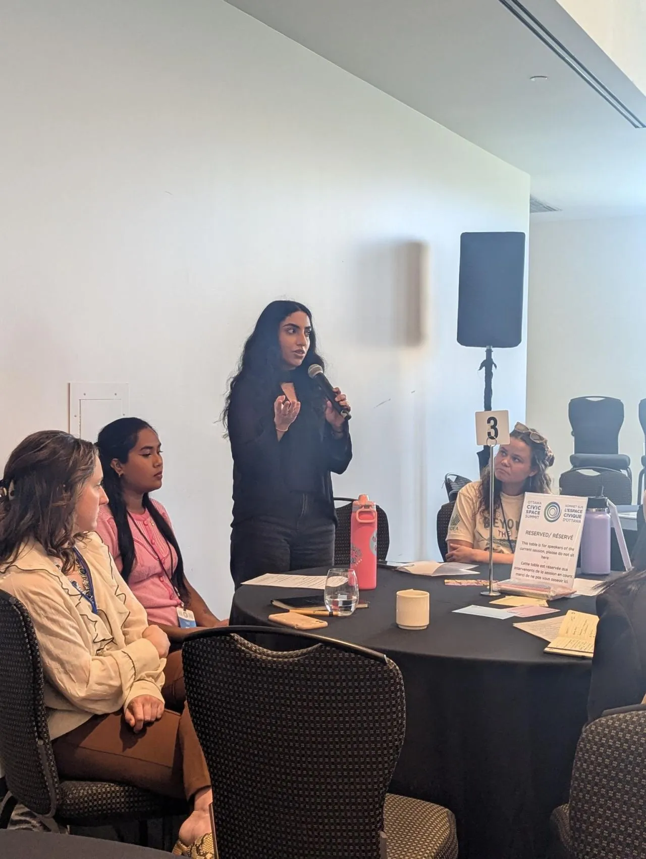 Mishika Khurana standing with a microphone, speaking to a roundtable of seated participants.